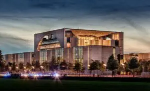 Ein modernes, beleuchtetes Gebäude mit einem markanten rechteckigen Design und großen Glasfenstern vor einem Abendhimmel. Das Gebäude ist von Bäumen umgeben und im Vordergrund sind Lichtstreifen vom Verkehr zu sehen.